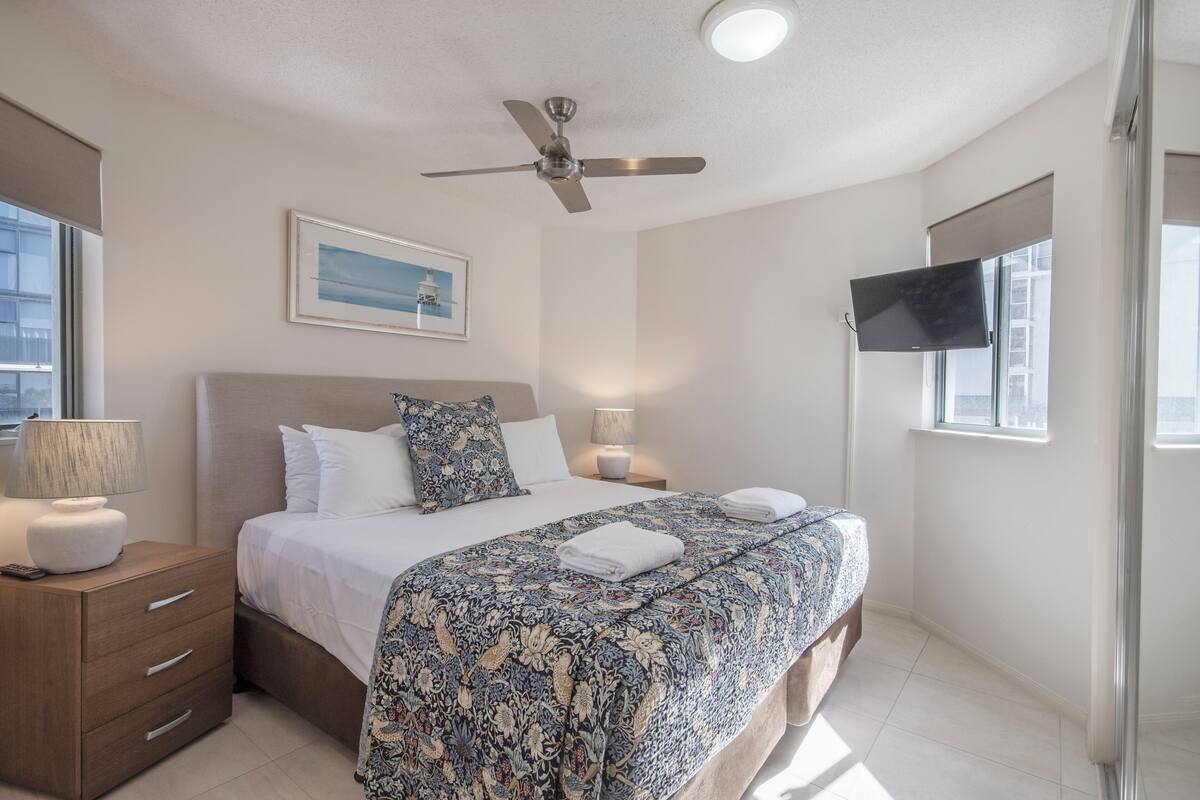 A modern bedroom with a bed, bedside lamps, a ceiling fan, a flat-screen TV, and a window.