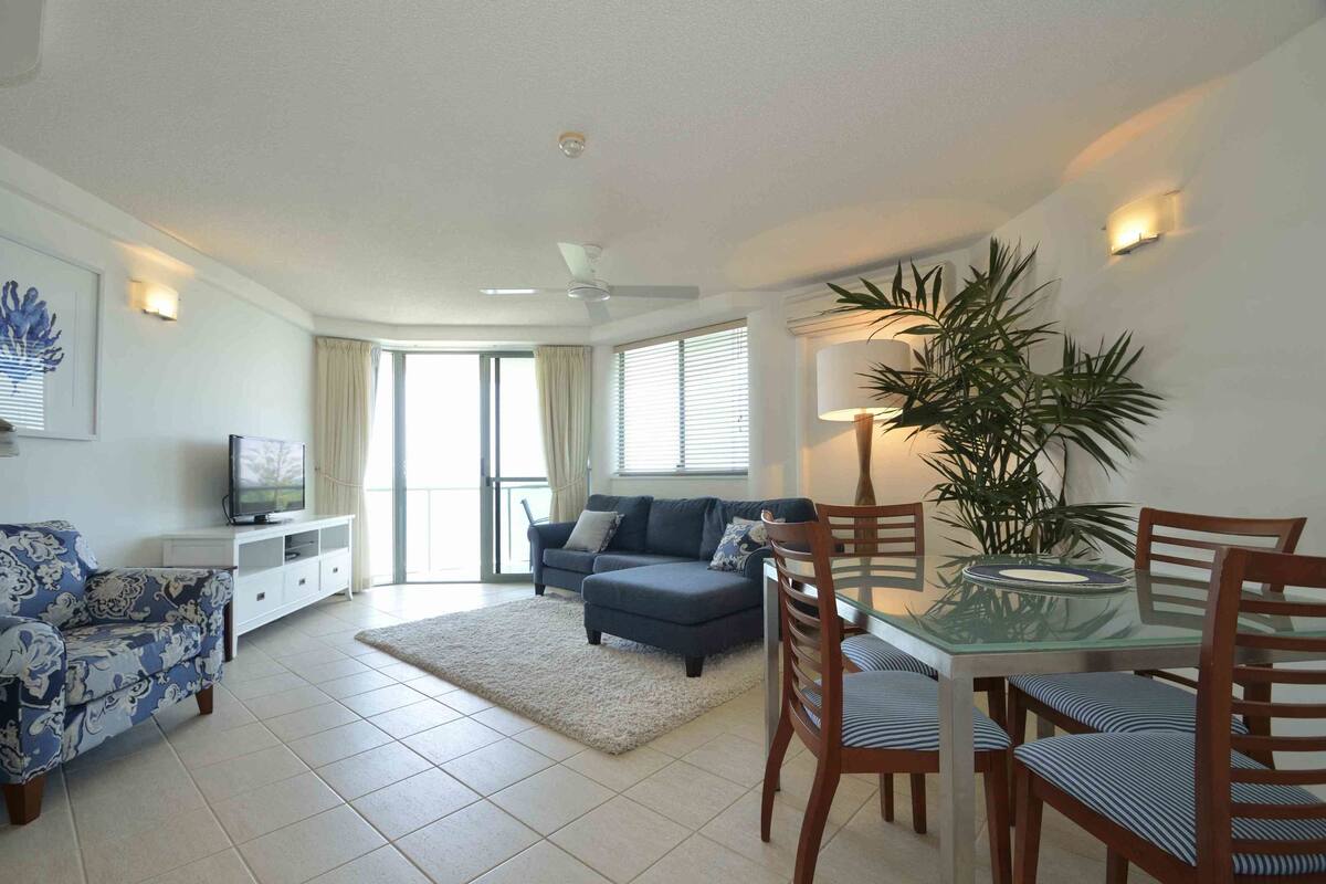 A living room with a sofa, a glass dining table with chairs, a TV on a stand, and a potted plant.