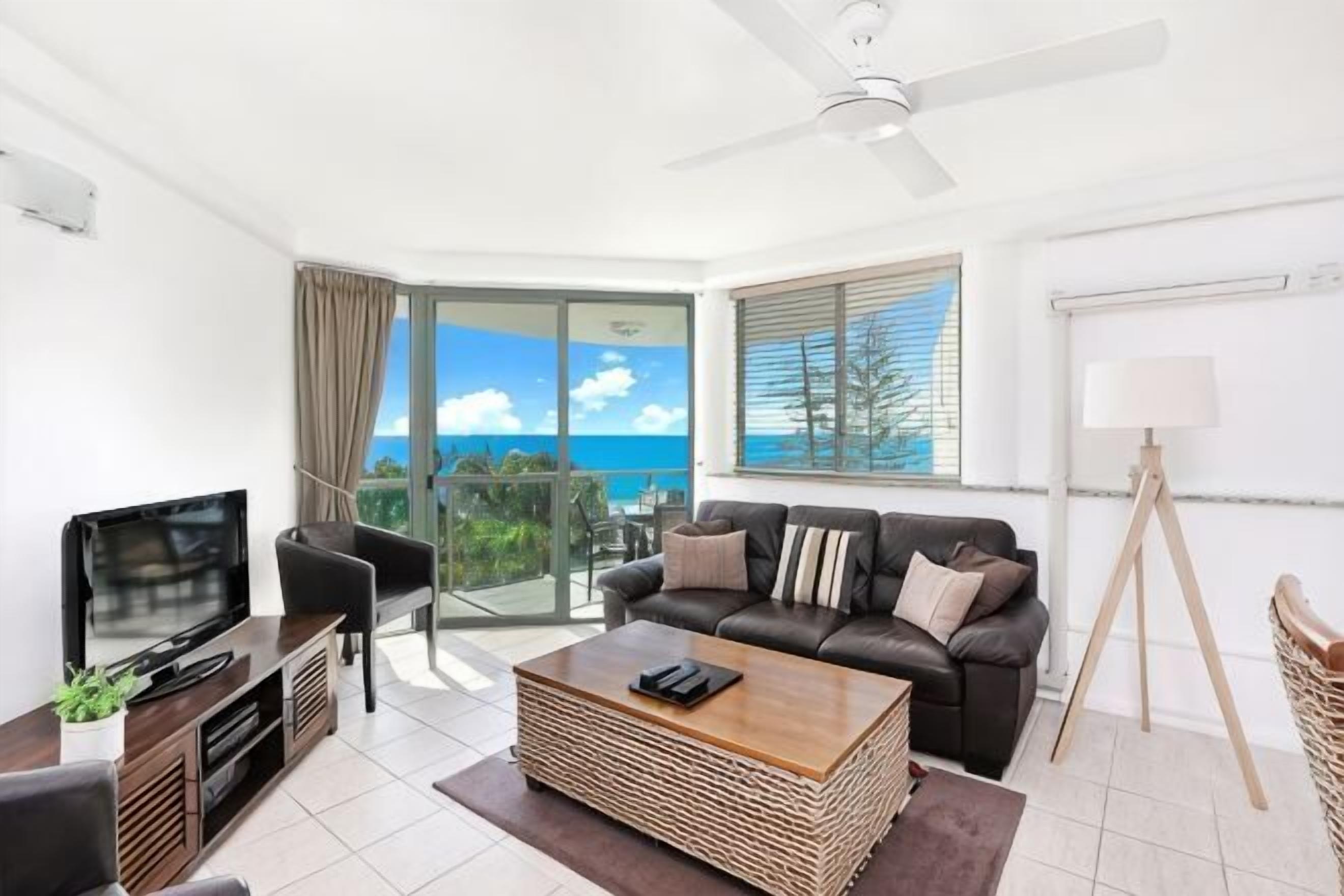 A modern living room with a flat-screen TV, a sofa, a coffee table, and a balcony with a view of the ocean.