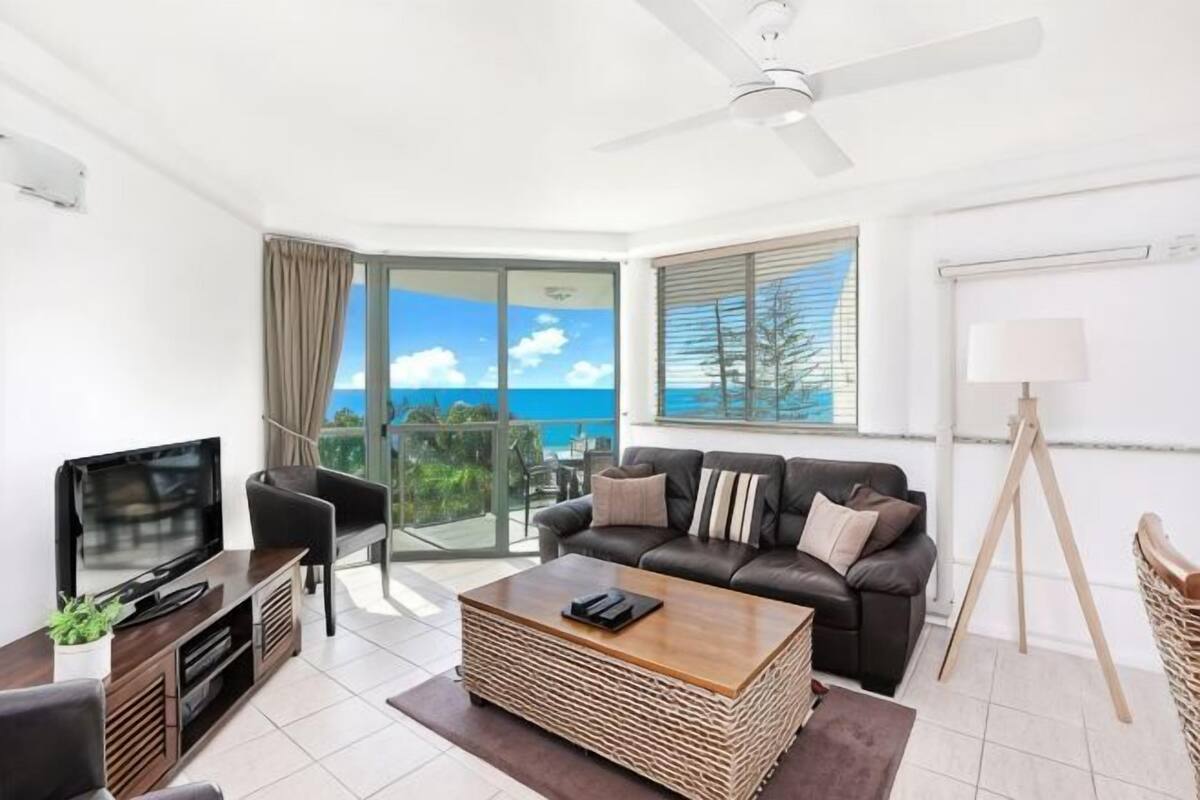 A modern living room with a flat-screen TV, a sofa, a coffee table, and a balcony with a view of the ocean.