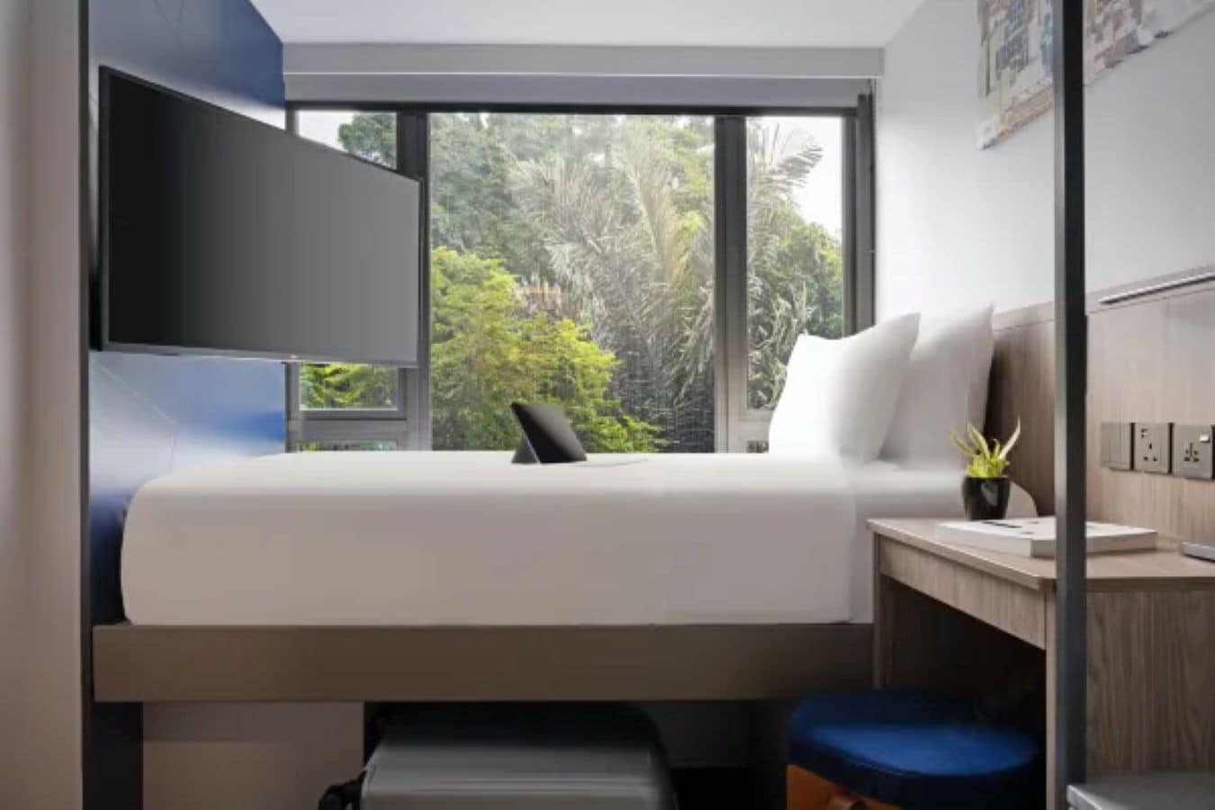 Ein modernes Zimmer mit Bett, Schreibtisch, Stuhl, Fenster mit Blick ins Grüne und einem Wand-Fernseher.
