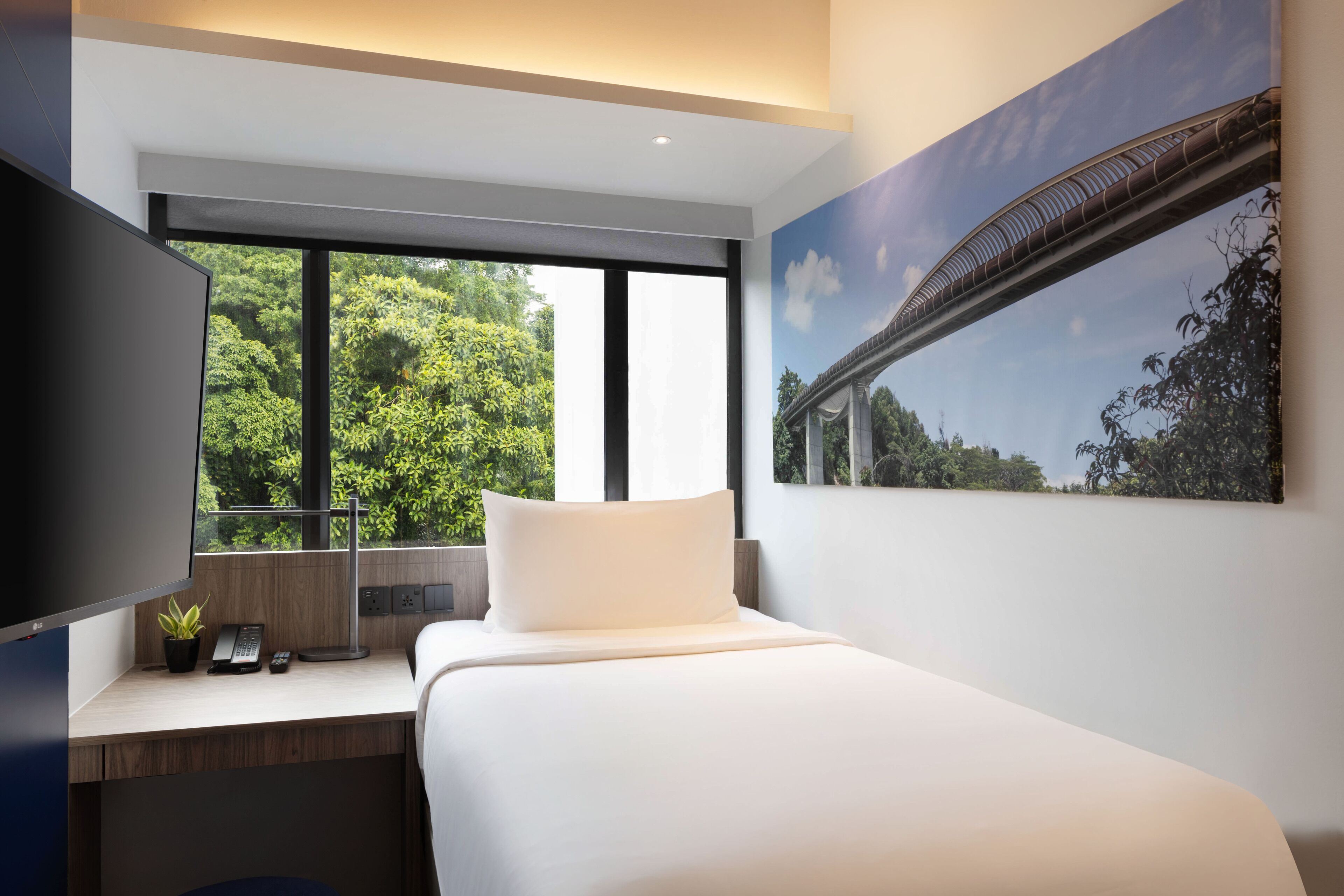 A modern hotel room with a large bed, a desk with a computer and phone, a TV, and a window with a view of trees and a bridge.