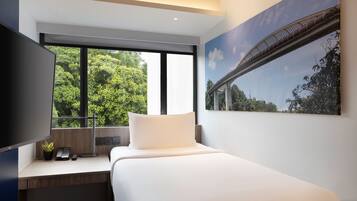A modern hotel room with a large bed, a desk with a computer and phone, a TV, and a window with a view of trees and a bridge.