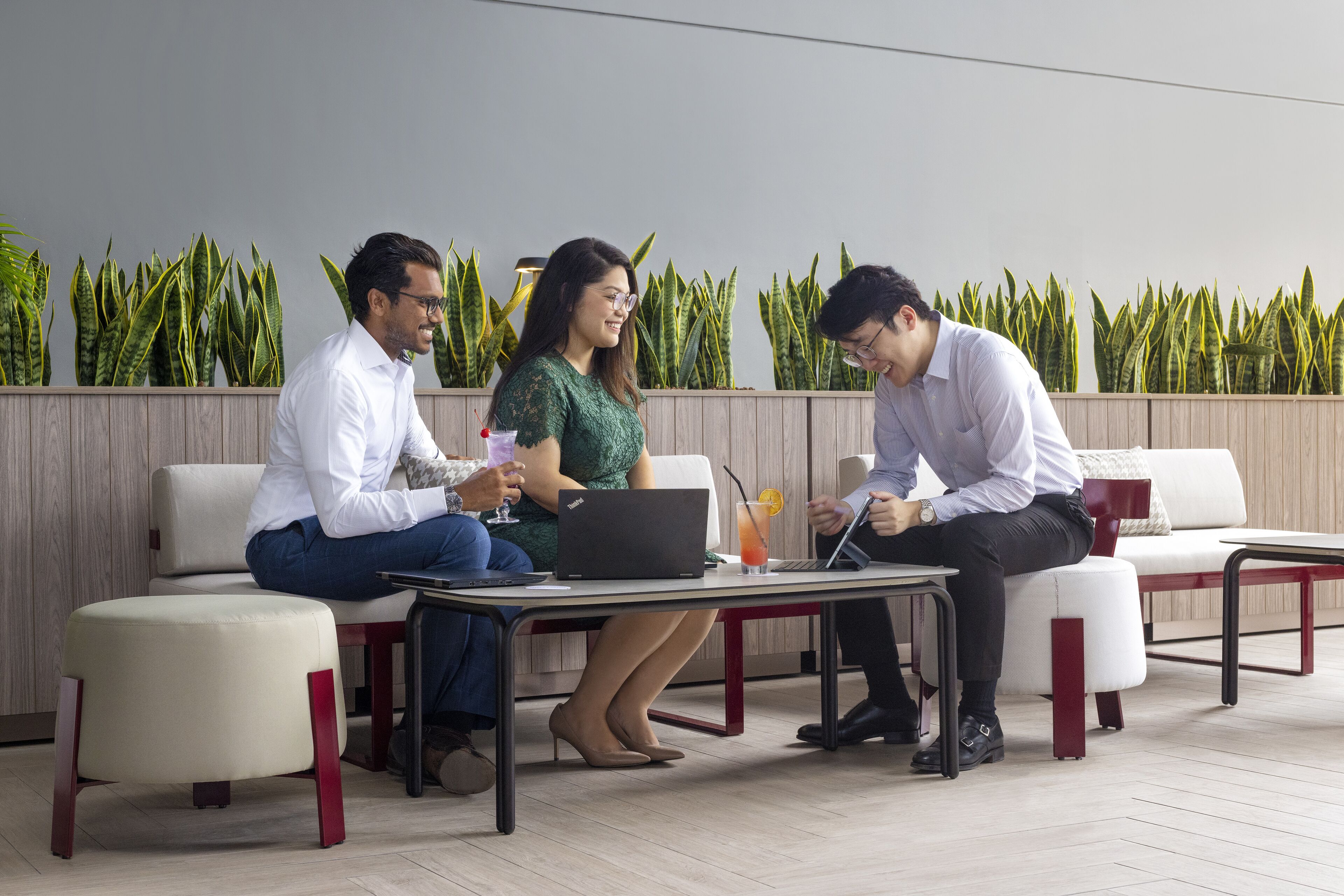 Three people seated at a table with laptops, in a modern outdoor setting with a wooden wall and green plants.