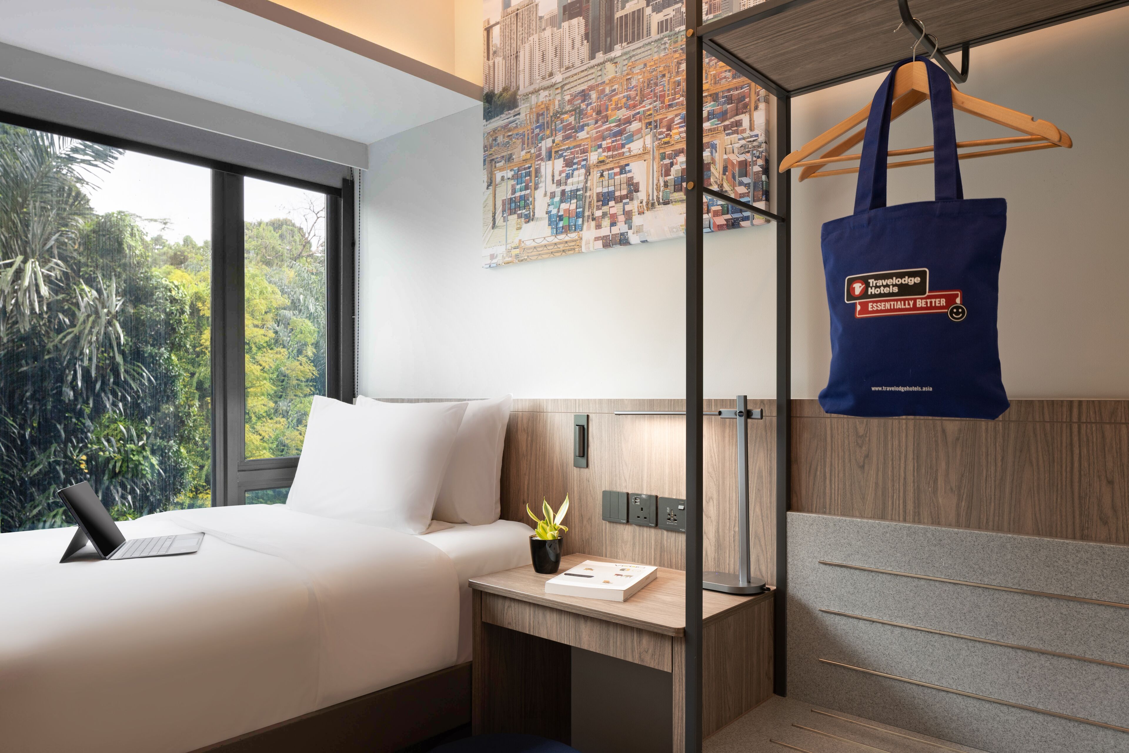 A hotel room with a bed, a desk, a laptop, a plant, and a wall with a poster.