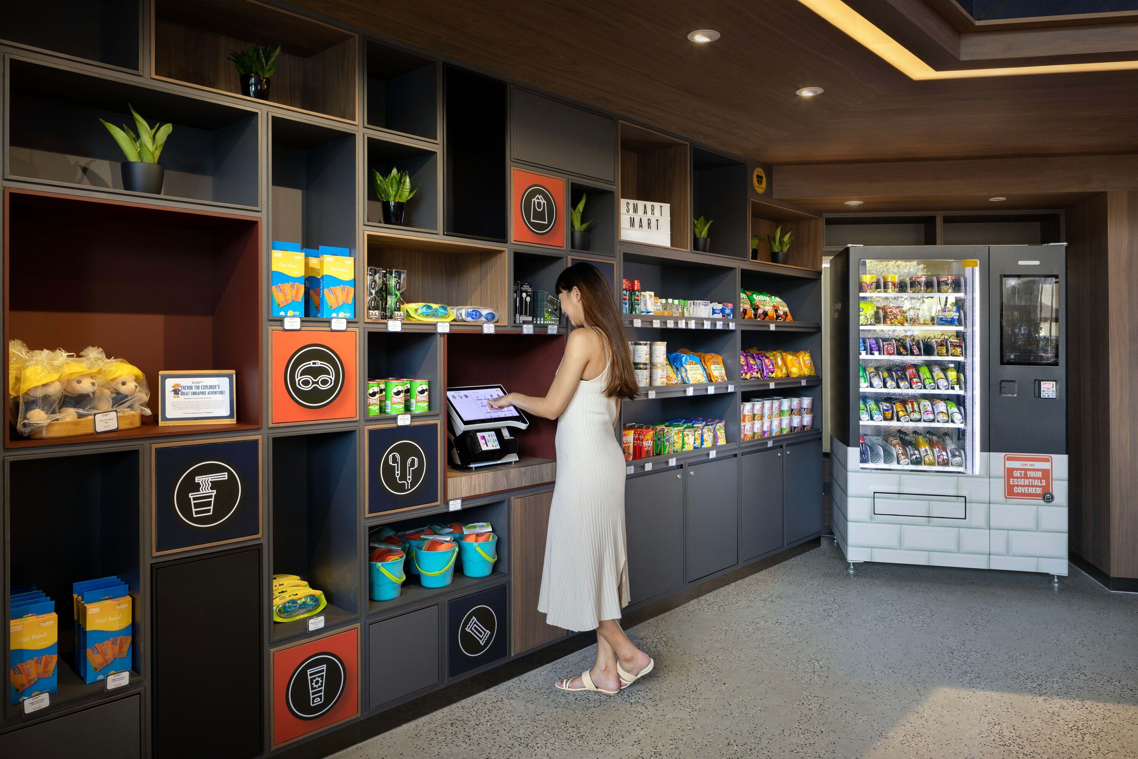 A woman at a checkout counter in a store with shelves stocked with snacks and a self-service kiosk.