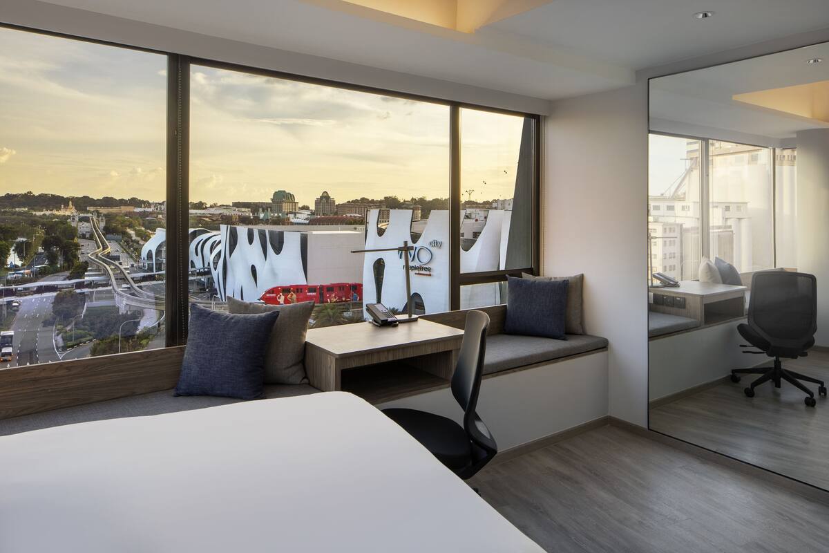 Ein modernes Hotelzimmer mit großem Fenster und Blick auf die Stadt, einem Schreibtisch mit Stuhl sowie einem Bett mit Ausblick.