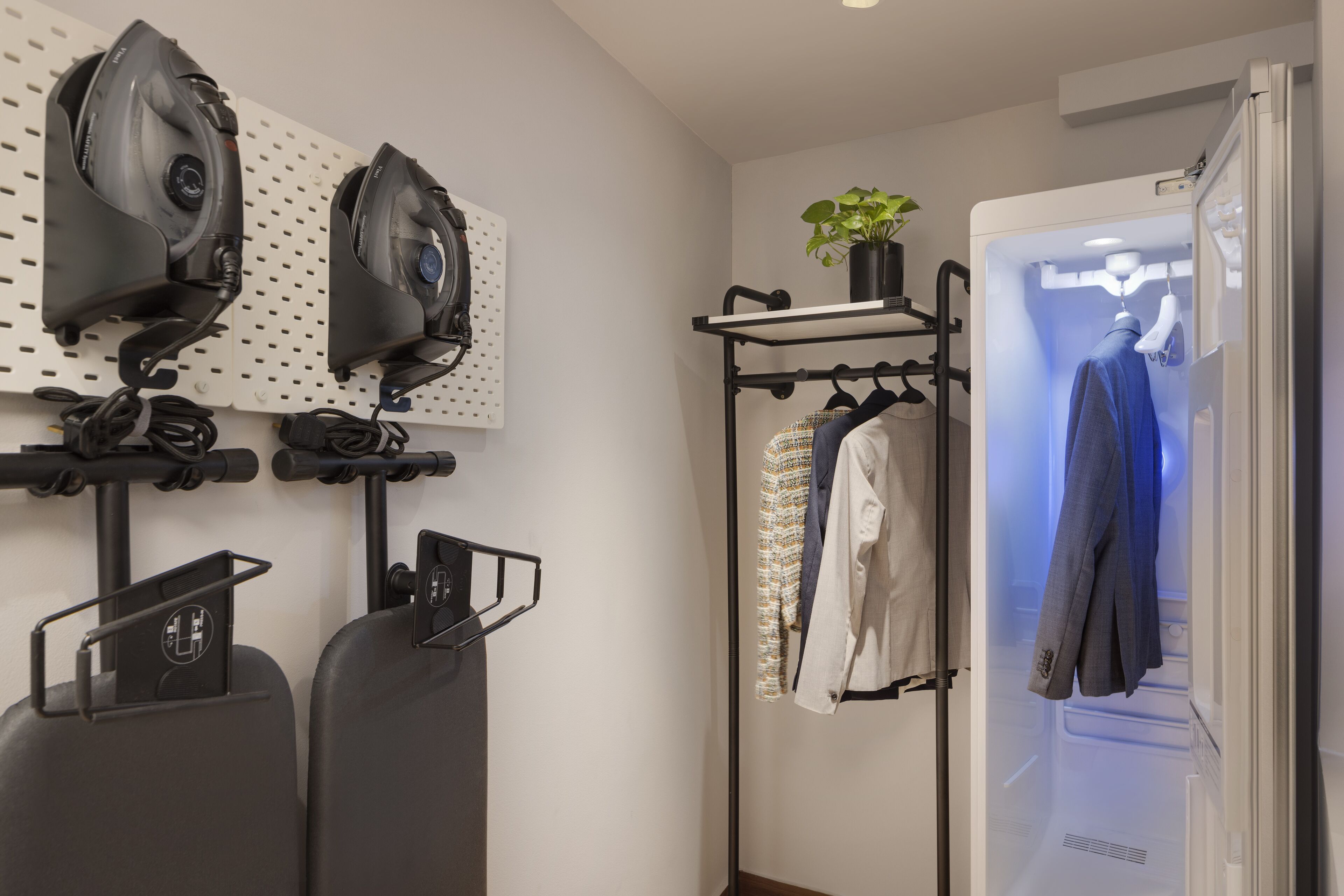 A room with two wall-mounted irons, a clothing rack with various garments, and a potted plant.