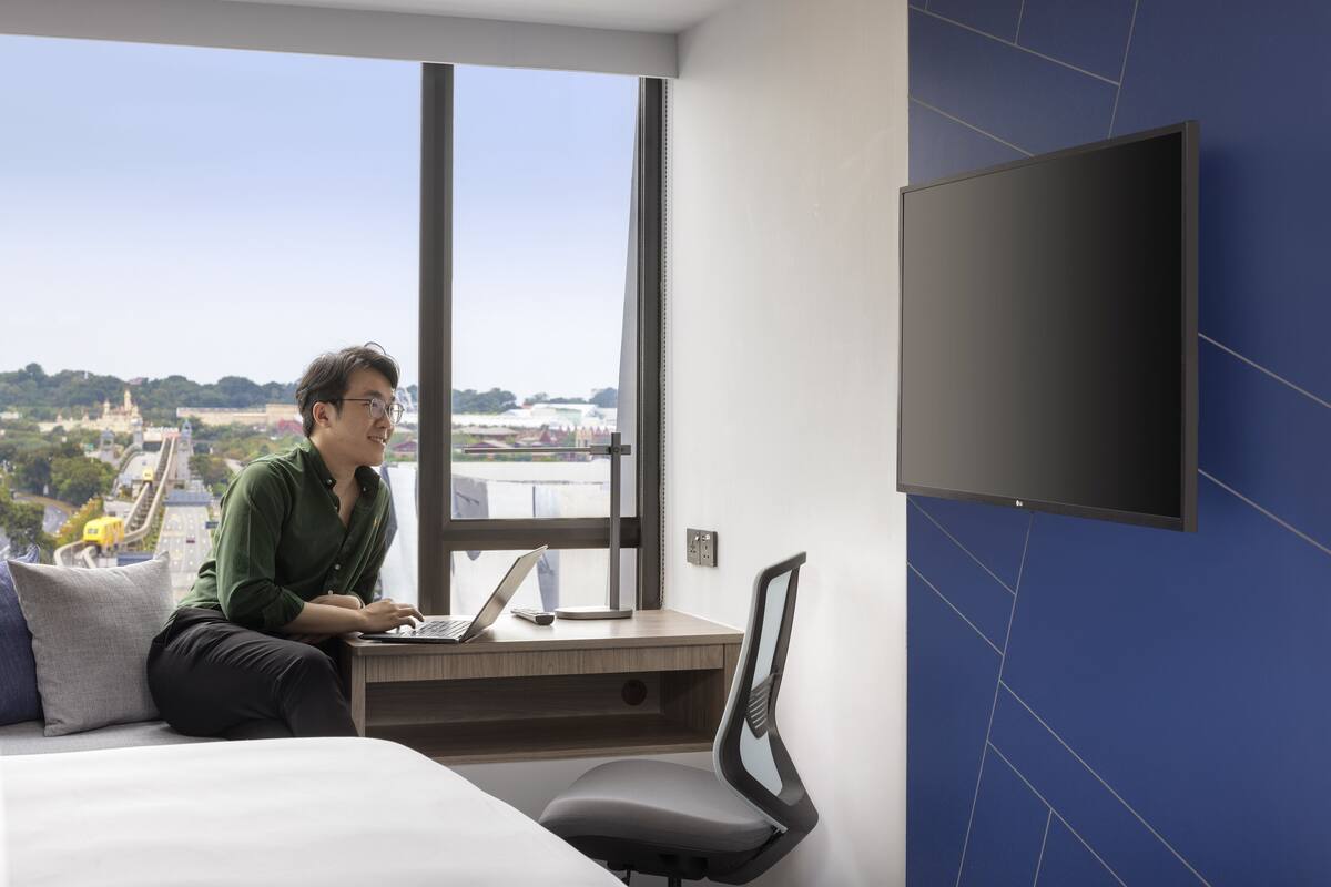 Ein modernes Hotelzimmer mit großem Fenster, einem wandmontierten Flachbildfernseher, einem Schreibtisch mit Laptop und einem bequemen Sessel.
