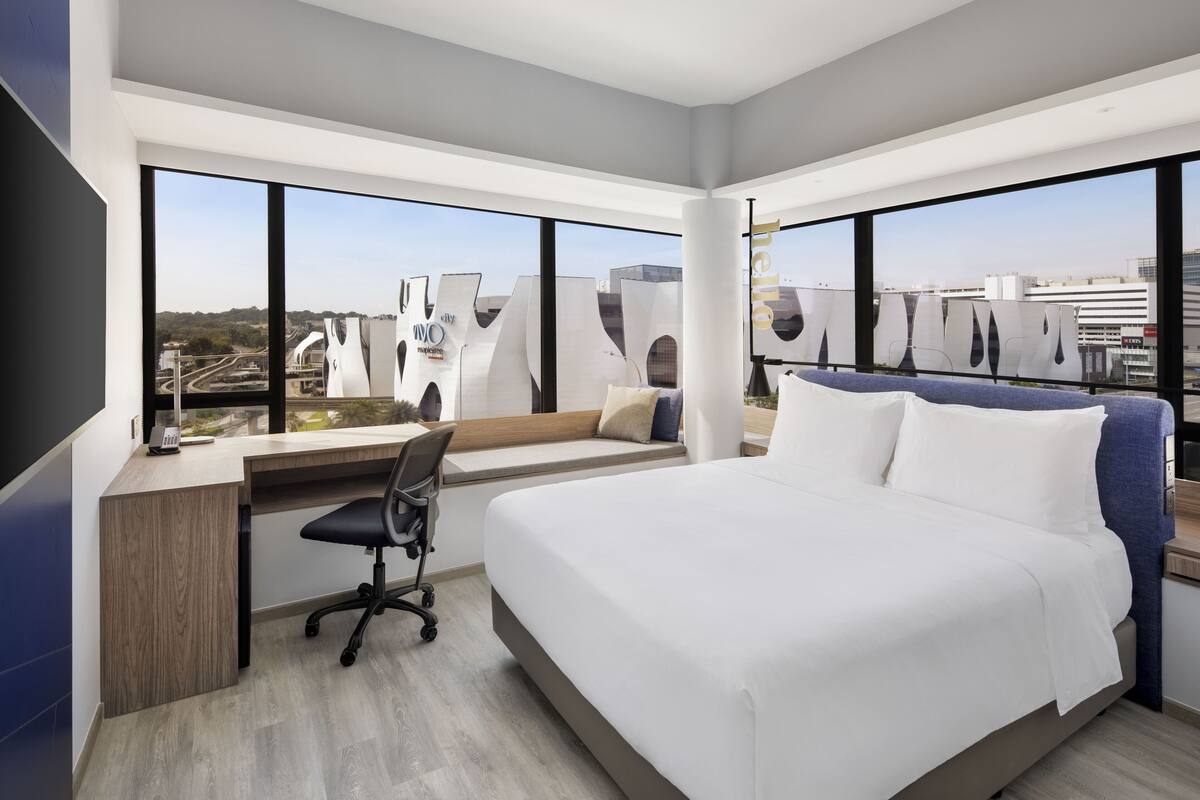 Ein modernes Hotelzimmer mit einem großen Bett, einem Schreibtisch mit Stuhl und Blick auf die Stadt durch große Fenster.