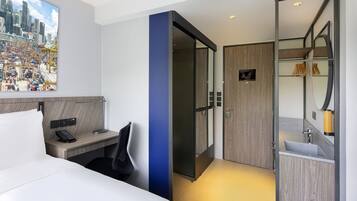 Et moderne hotelværelse med seng, skrivebord med telefon, spejl, badeværelse med håndvask og udsigt over bybilledet.