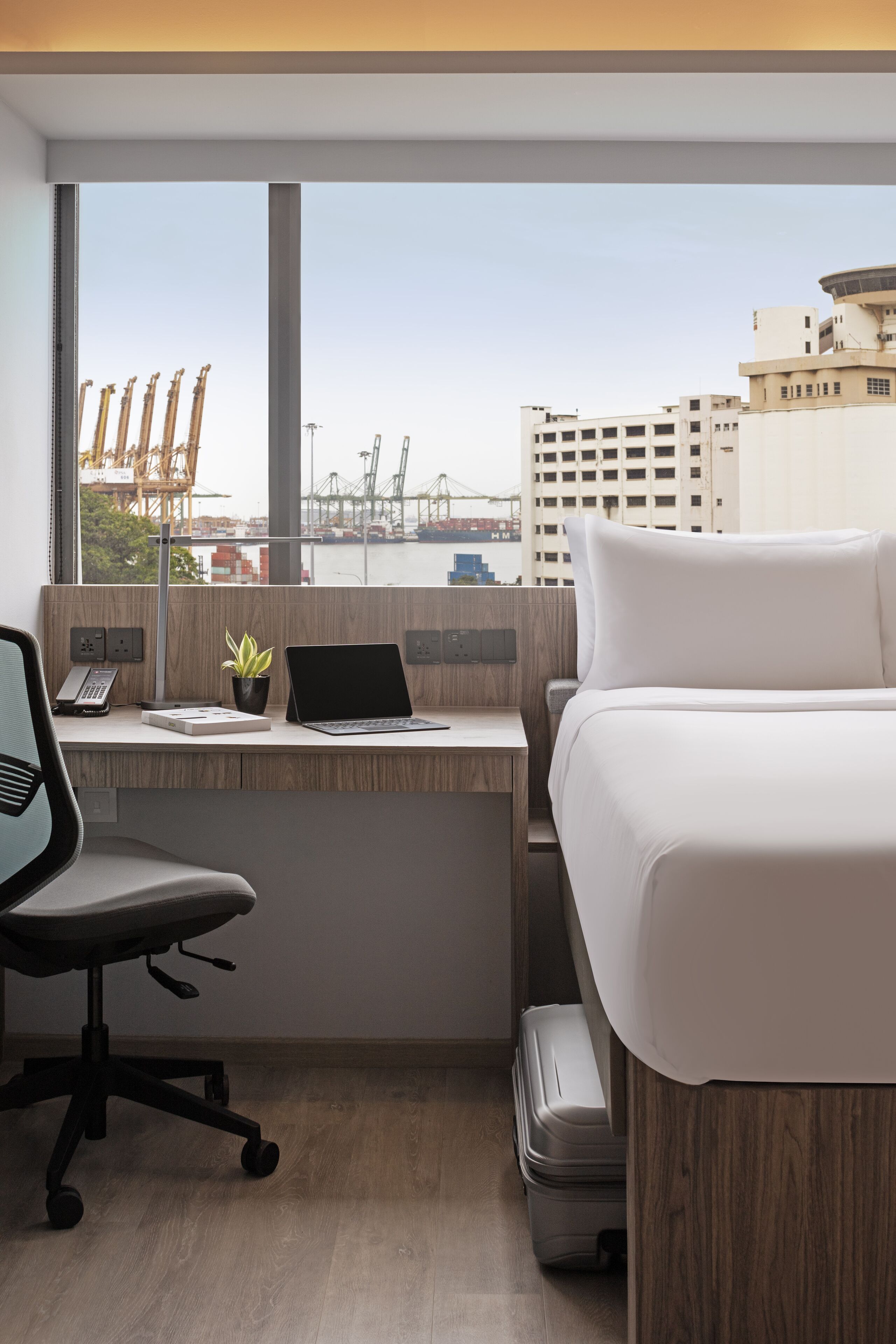 Habitación de hotel con escritorio, ordenador portátil, silla, cama y vistas a la ciudad y al puerto.