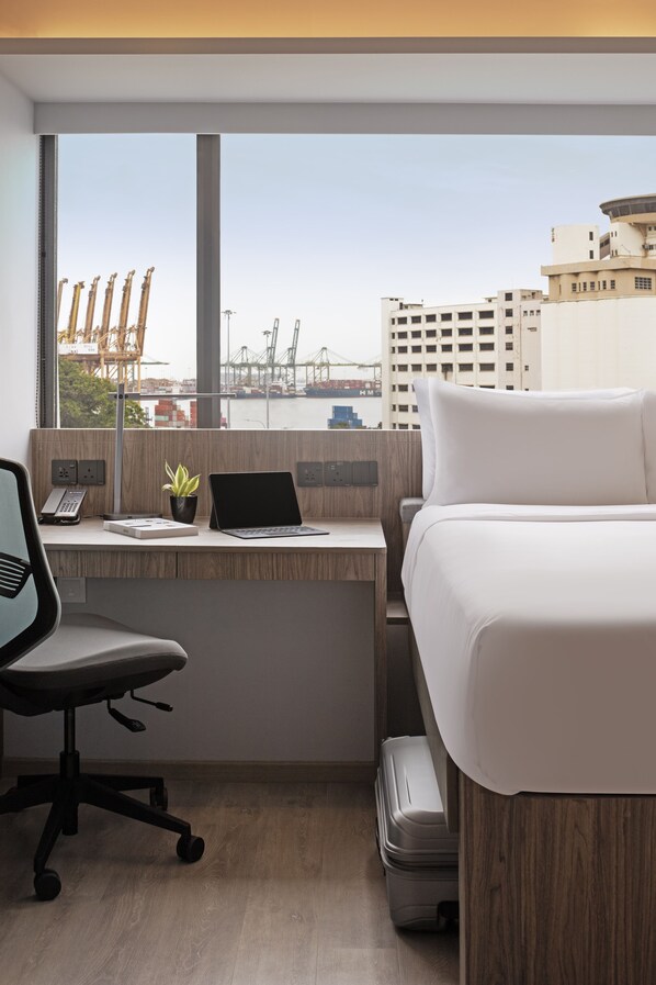 Habitación de hotel con escritorio, ordenador portátil, silla, cama y vistas a la ciudad y al puerto.