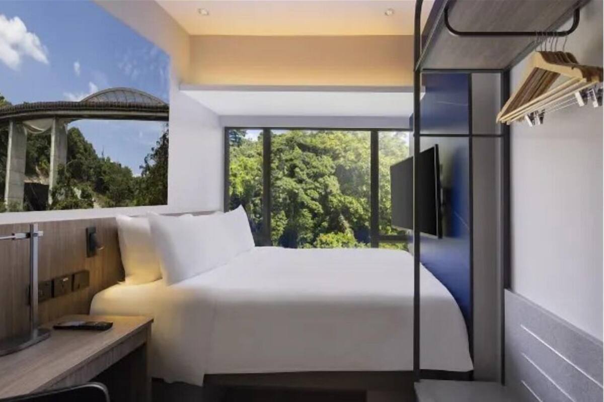 Ein modernes Hotelzimmer mit Bett, Schreibtisch, Fernseher und einem großen Fenster mit Blick auf Bäume.