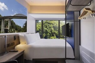 Quarto de hotel moderno com cama, escrivaninha, TV e uma janela grande com vista para árvores.