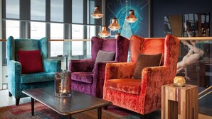 A modern lounge area with a teal armchair, a purple armchair, and an orange armchair, each with a cushion. A wooden coffee table with a glass candle holder is in the center. - Scandic Vulkan (Oslo)