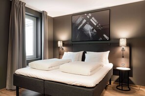 A neatly made bed with white linens, a dark headboard, and a large abstract painting above it. There are two wall-mounted lamps on either side of the bed.