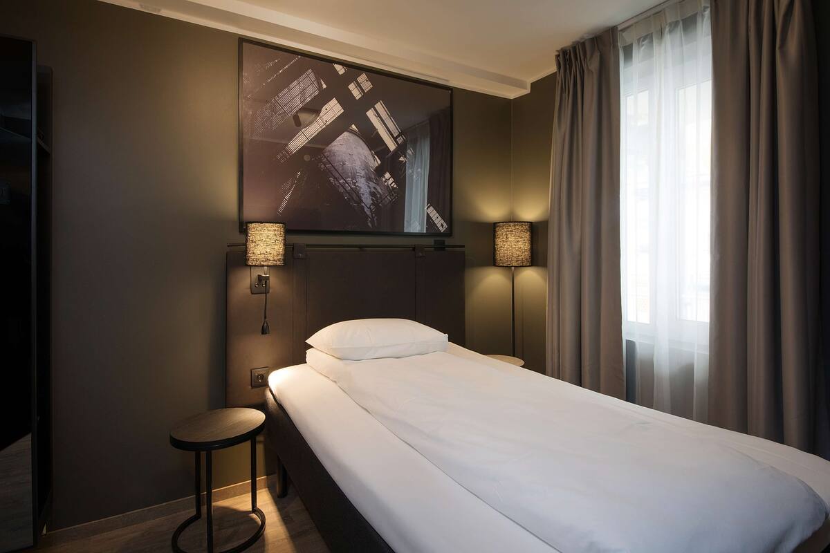 Habitació d'hotel amb un llit, un televisor gran penjat a la paret, dues làmpades de nit i una finestra amb cortines.