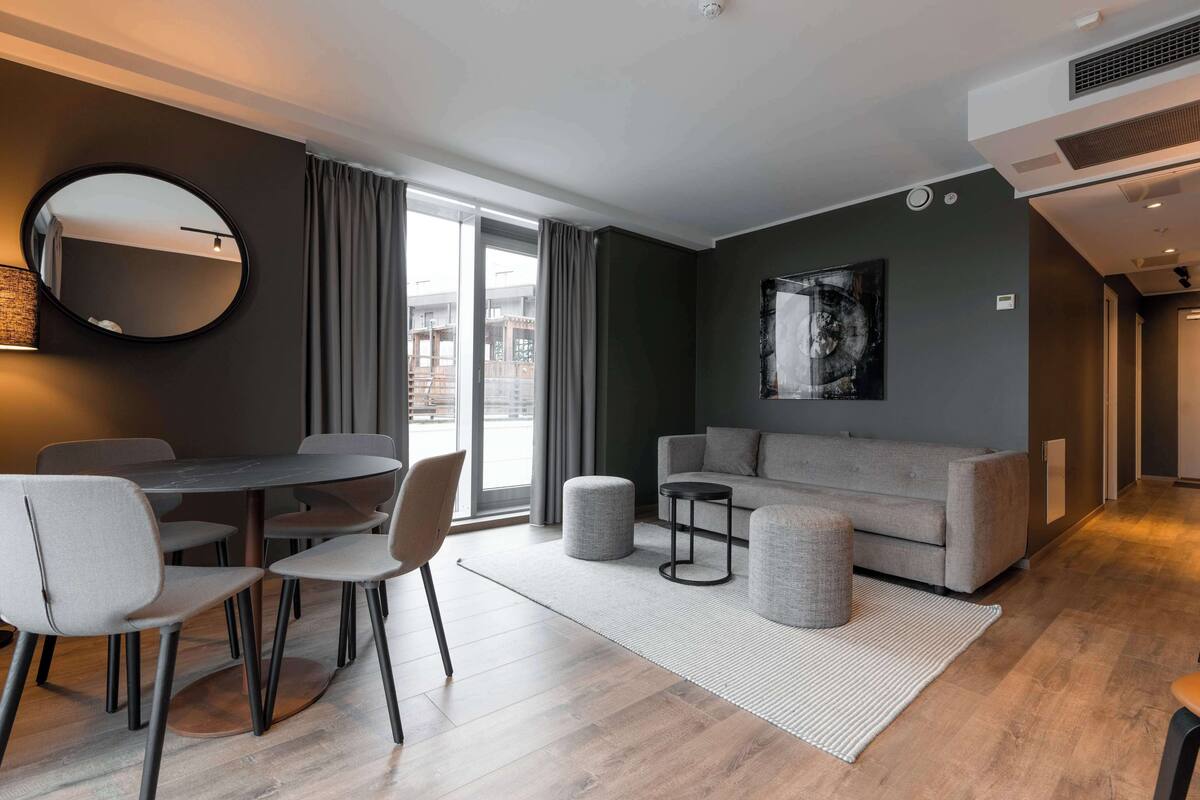 En moderne stue med en grå sofa, et rundt bord med stole, et rundt spejl og et stort vindue med gardiner.