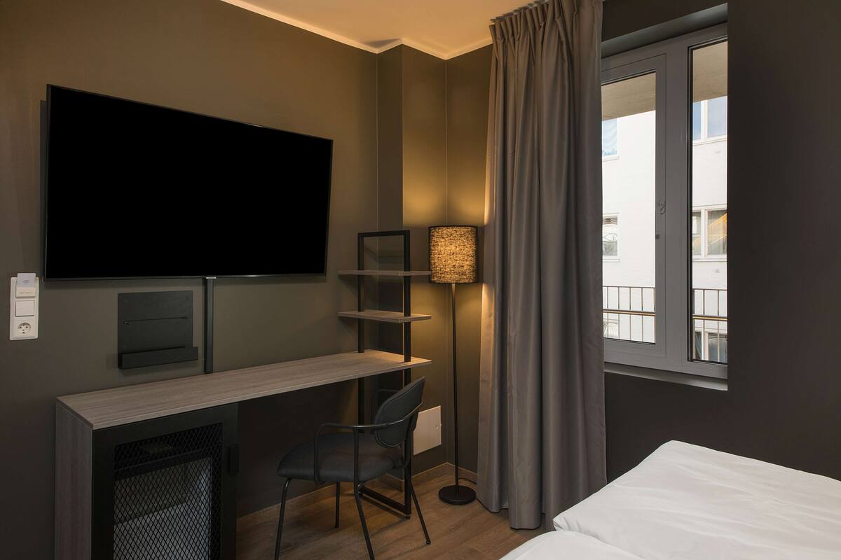 Habitació d'hotel amb llit, escriptori, cadira, televisió, làmpada i finestra amb cortines.