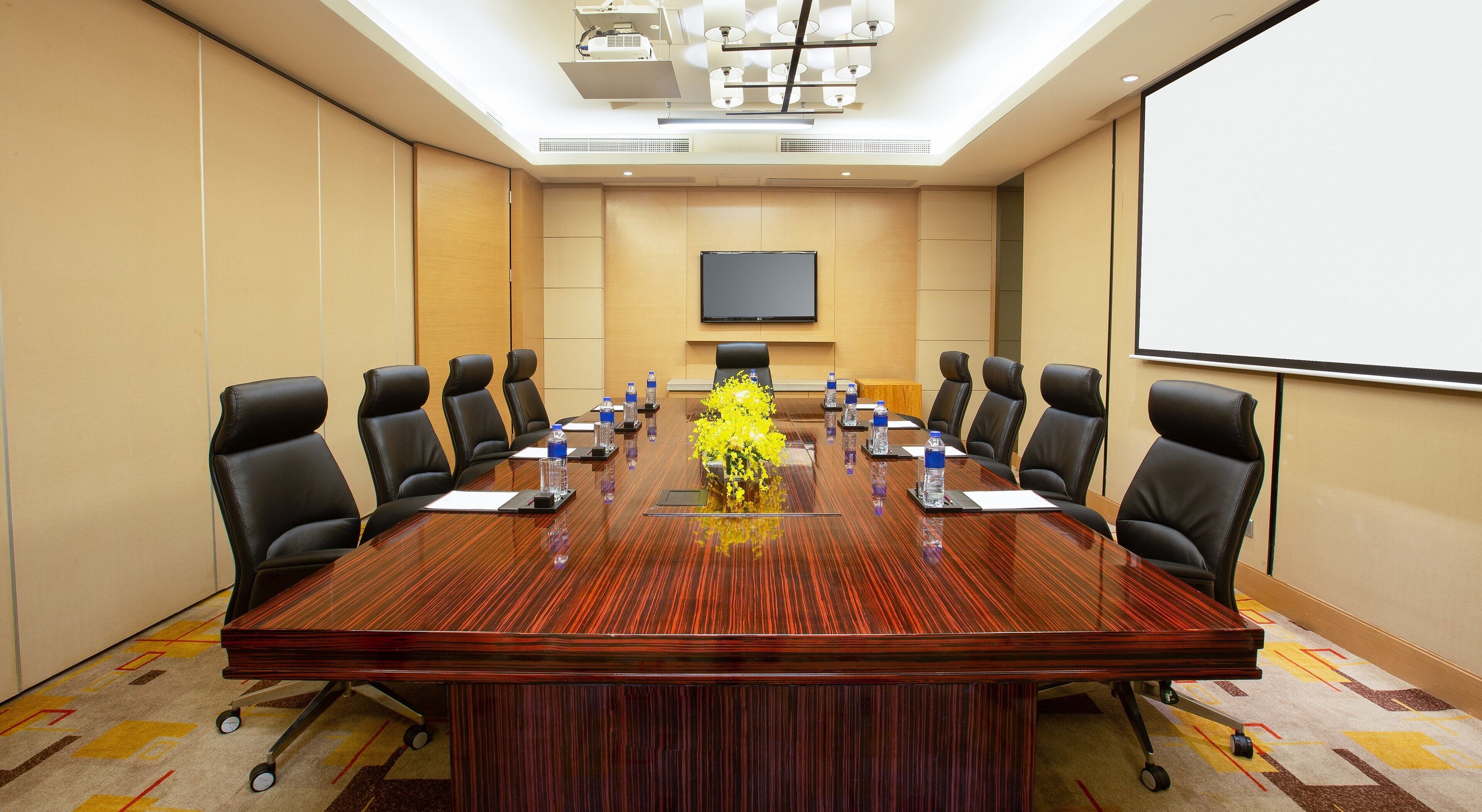 A conference room with a large wooden table, black leather chairs, and a projector screen on the wall.