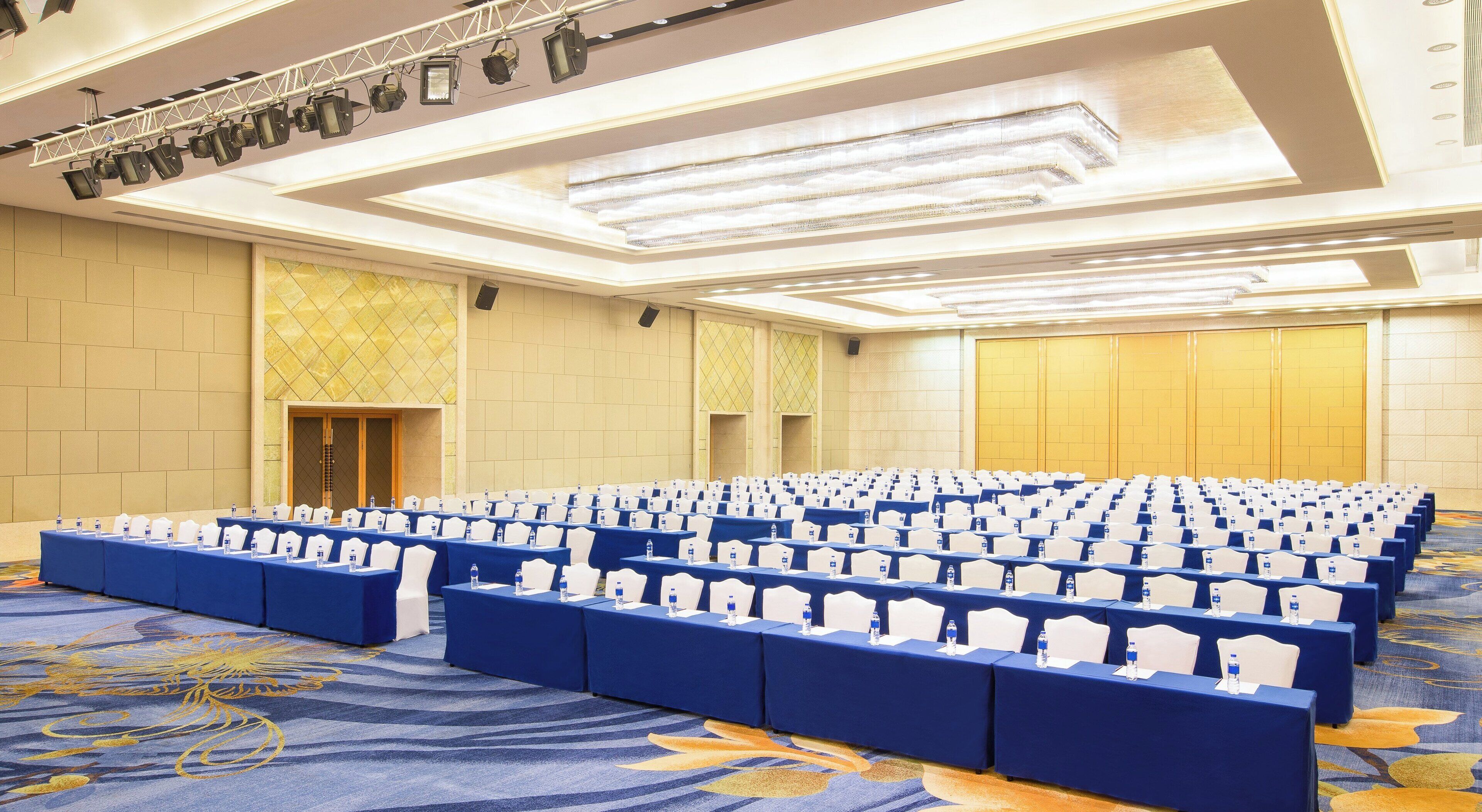 A conference room with rows of tables and chairs, a stage with lighting equipment, and a carpeted floor.