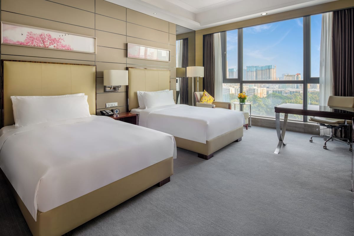 A hotel room with two beds, a desk, a chair, a lamp, and a view of the city through large windows.