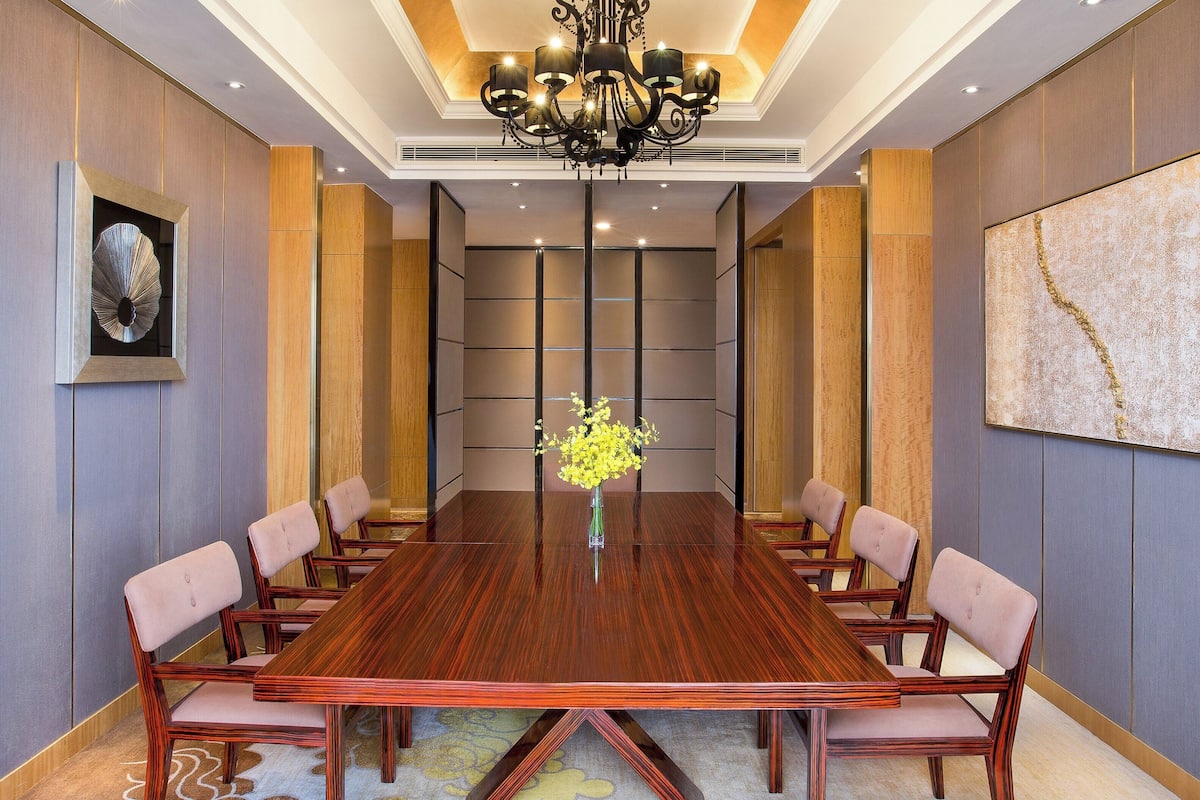 A conference room with a long wooden table, cushioned chairs, a chandelier, and a vase with yellow flowers.