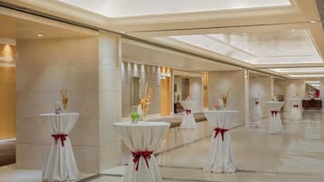 A spacious hotel lobby with multiple round tables, each adorned with a white tablecloth and a red ribbon. The lobby features a polished marble floor and a high ceiling with skylights.