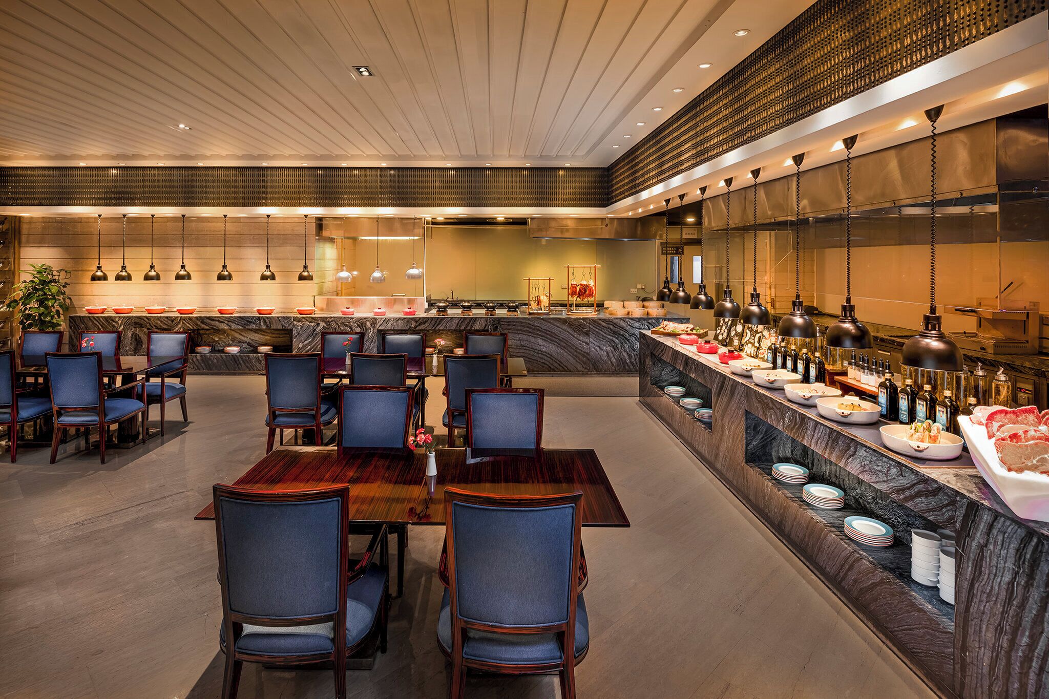 A spacious dining area with multiple tables and chairs, a marble counter with food and beverages, and pendant lighting.