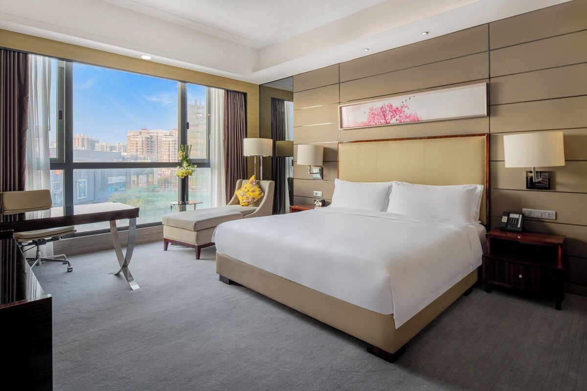 A hotel room with a large bed, a desk, a chair, and a view of the city through large windows.