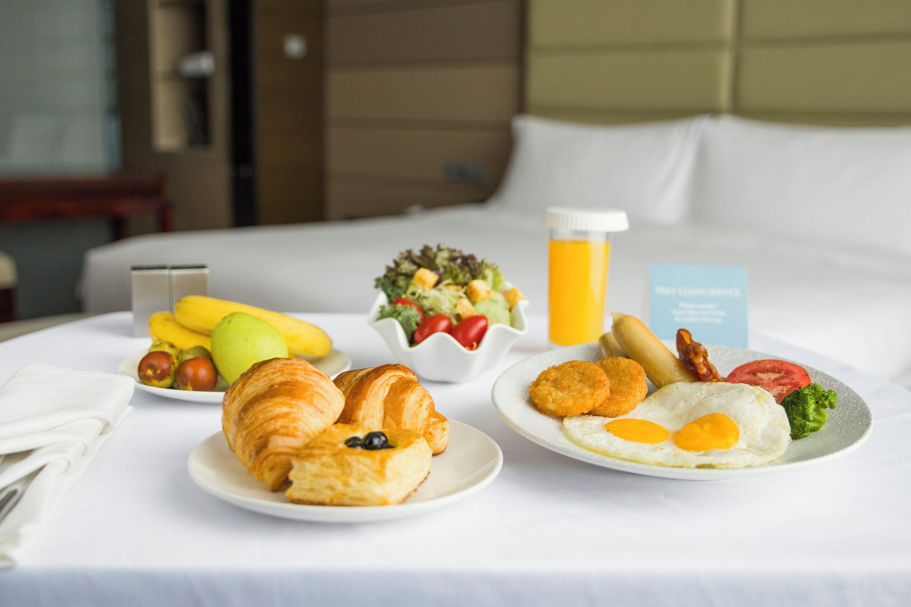 A breakfast setup on a hotel bed with croissants, eggs, fruit, and a salad.