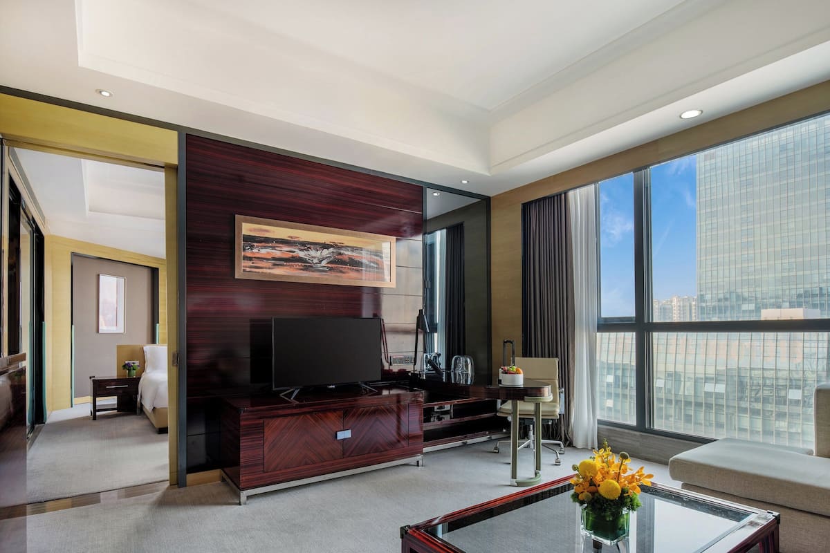 A modern hotel room with a large window, a flat-screen TV, a sofa, a coffee table with a vase, and a bed with a nightstand.