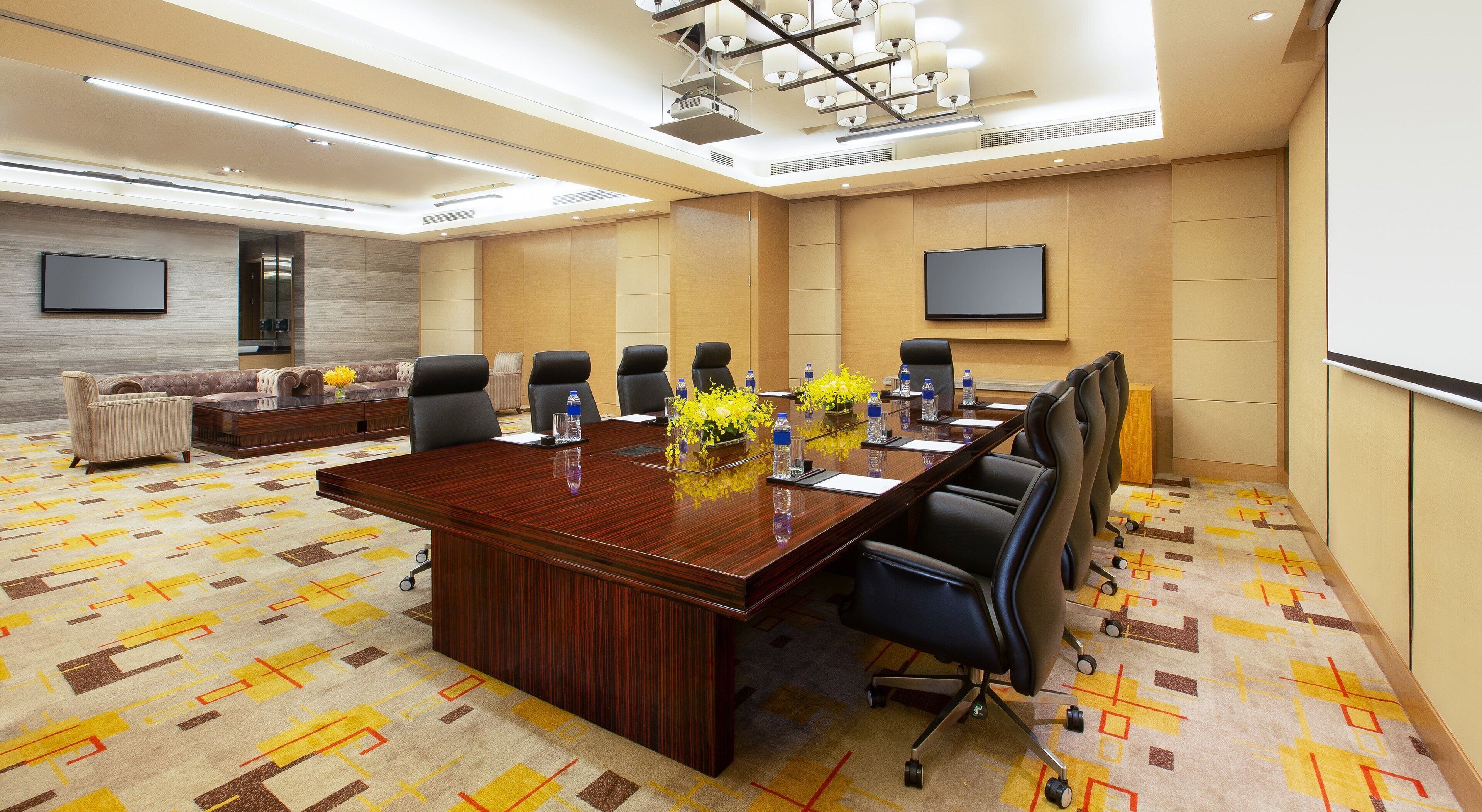 A conference room with a large wooden table, black chairs, and a sofa. There are two flat-screen TVs on the walls, a carpeted floor, and a chandelier.