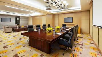 A conference room with a large wooden table, black chairs, and a sofa. There are two flat-screen TVs on the walls, a carpeted floor, and a chandelier.