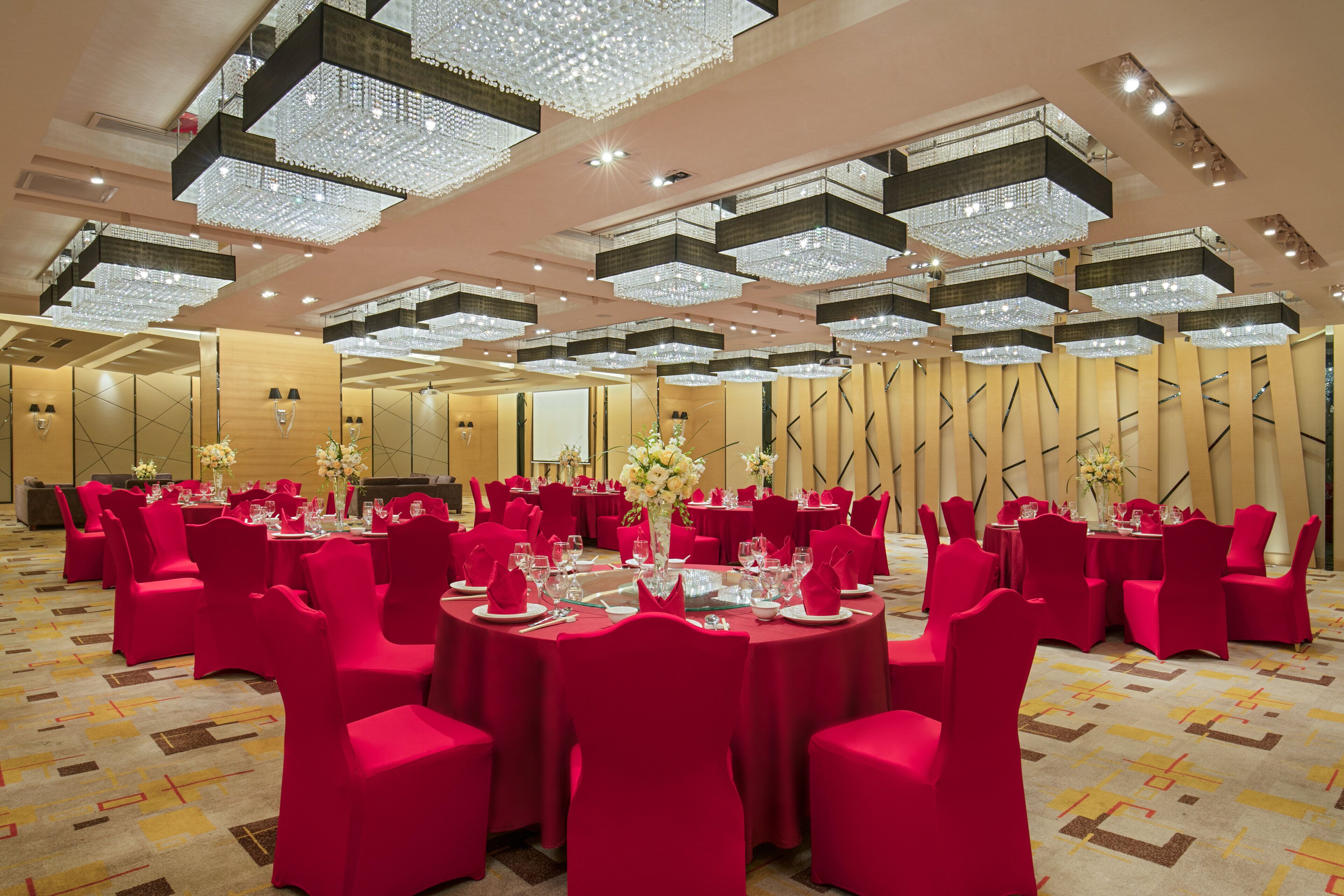 A banquet hall with round tables, red chairs, and chandeliers.