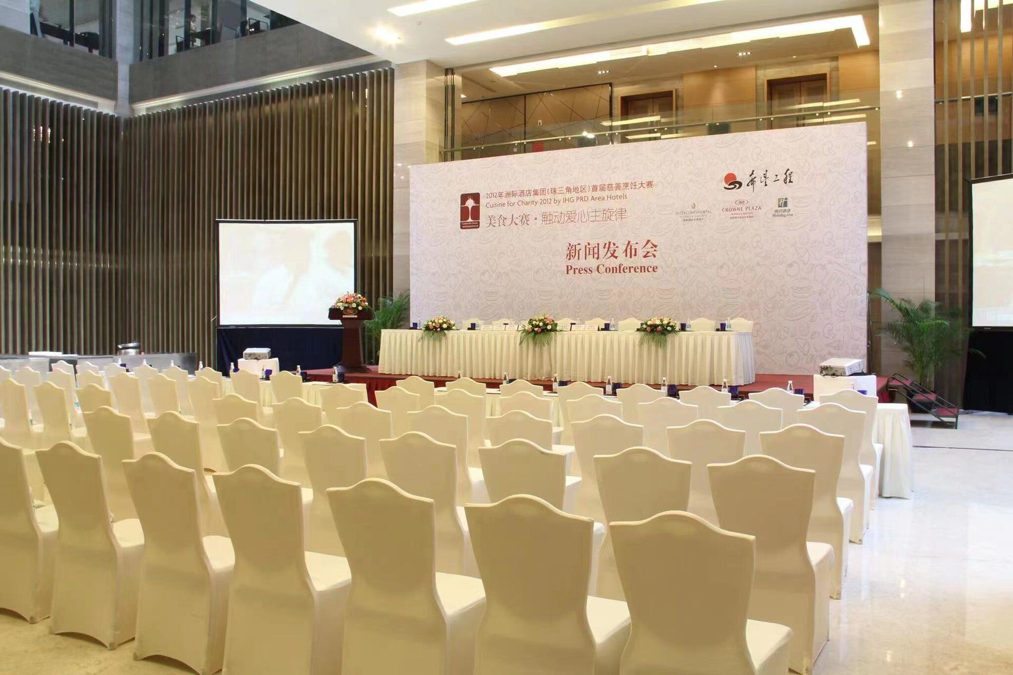 A conference room with rows of white chairs, a stage with a banner, and a large screen.