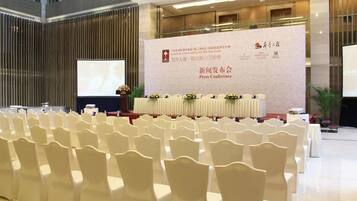 A conference room with rows of white chairs, a stage with a banner, and a large screen.