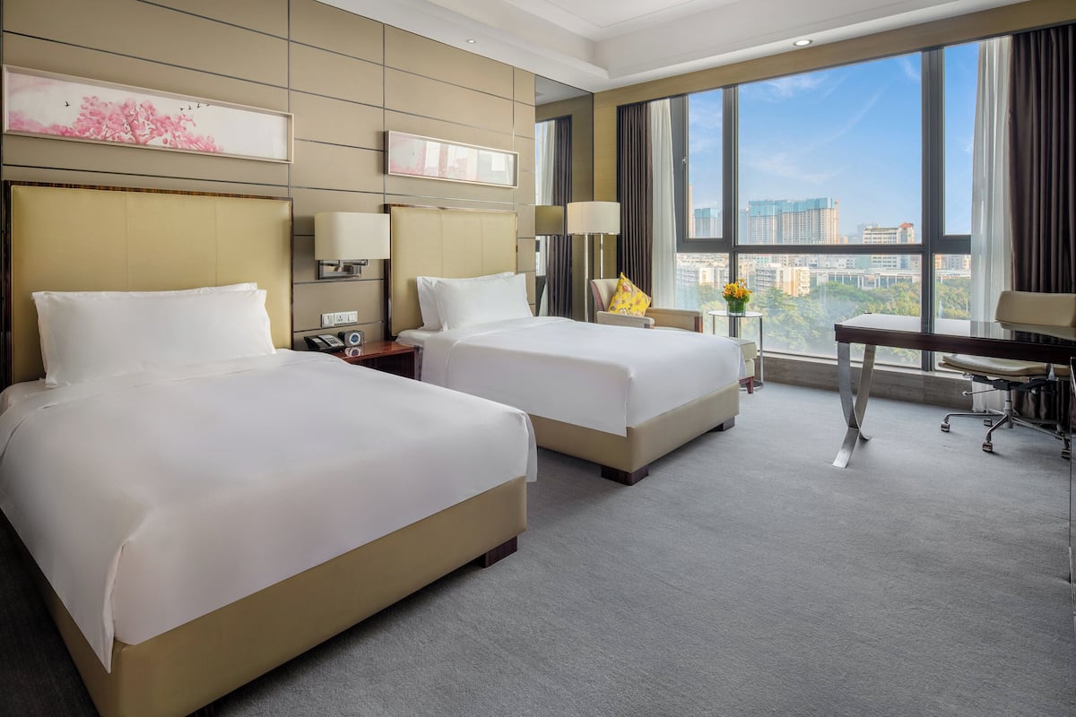 A hotel room with two beds, a desk, a chair, and a large window offering a city view.