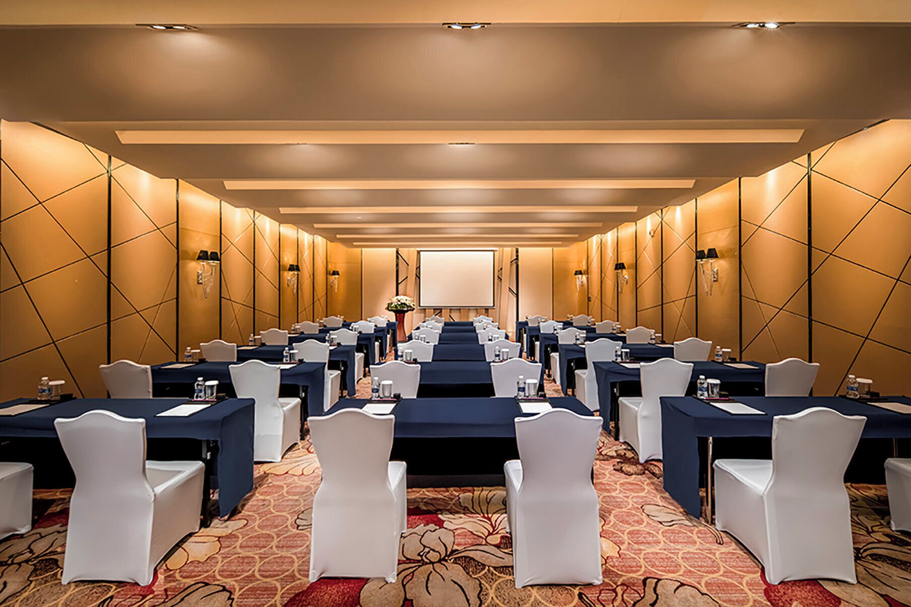 Sala de conferência com fileiras de mesas e cadeiras, equipada com garrafas de água e porta-nomes.