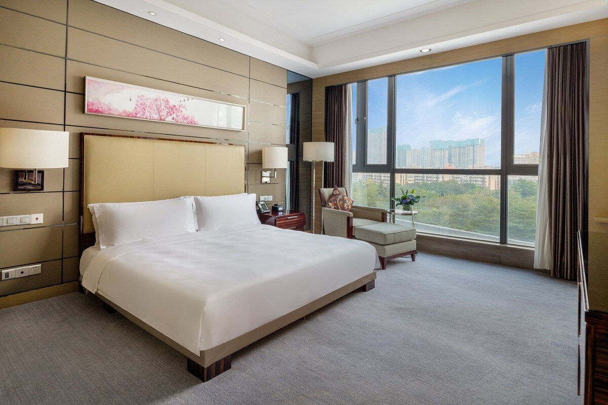 A spacious hotel room with a large bed, a desk with a chair, a view of the city through large windows, and a painting on the wall.