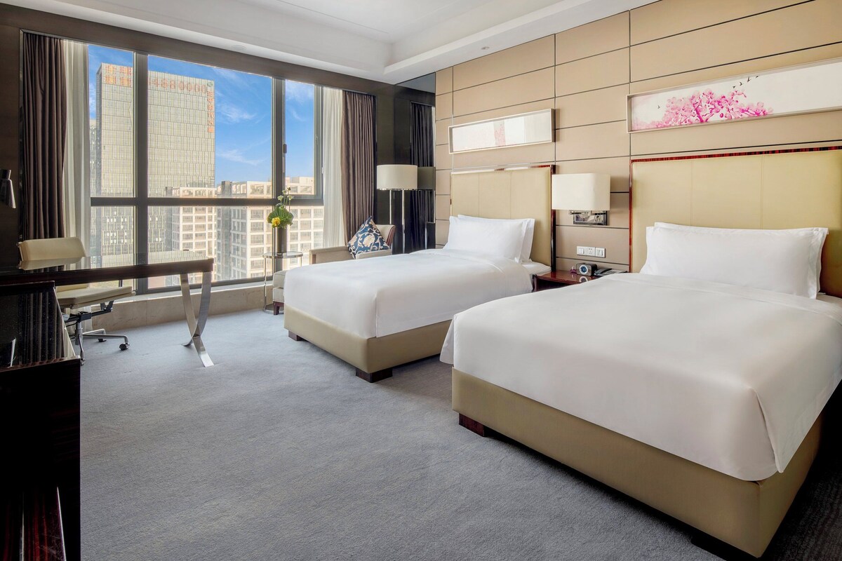 A hotel room with two beds, a desk, and a large window offering a city view.