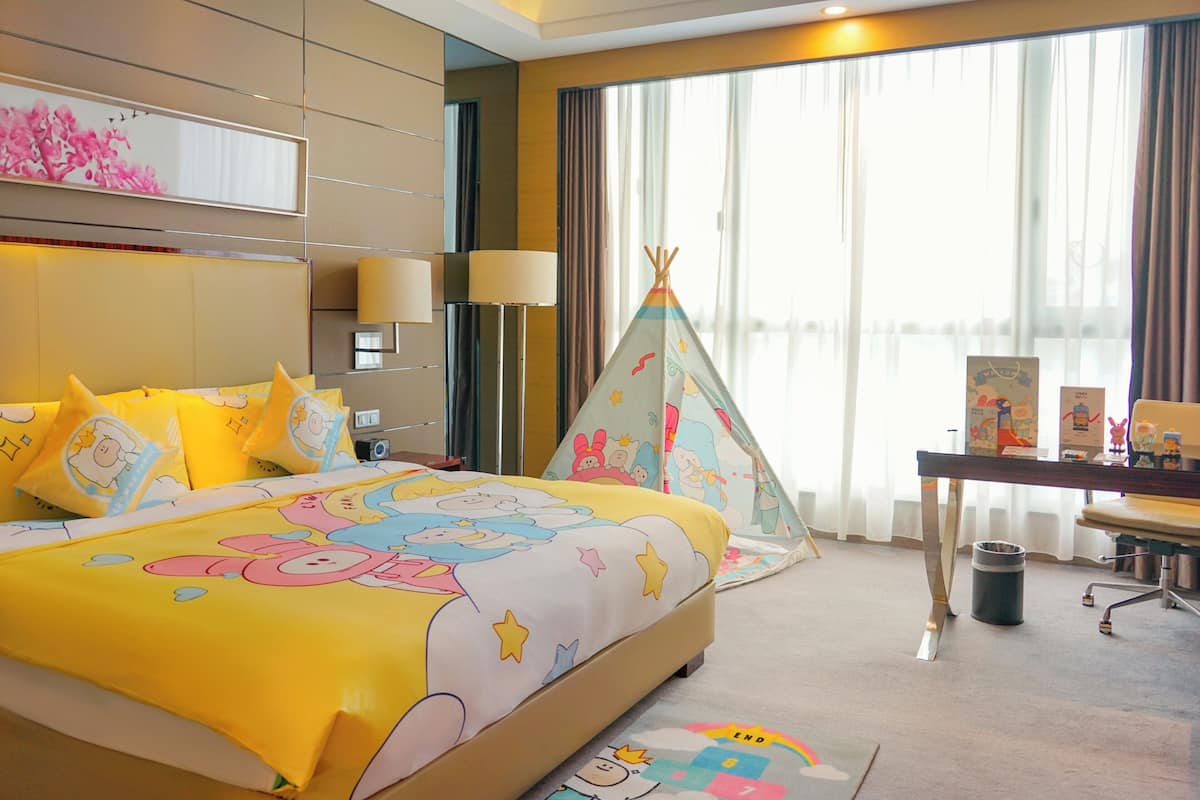A hotel room with a bed featuring a cartoon character design, a desk with a lamp, a window with curtains, and a small tent-like structure.