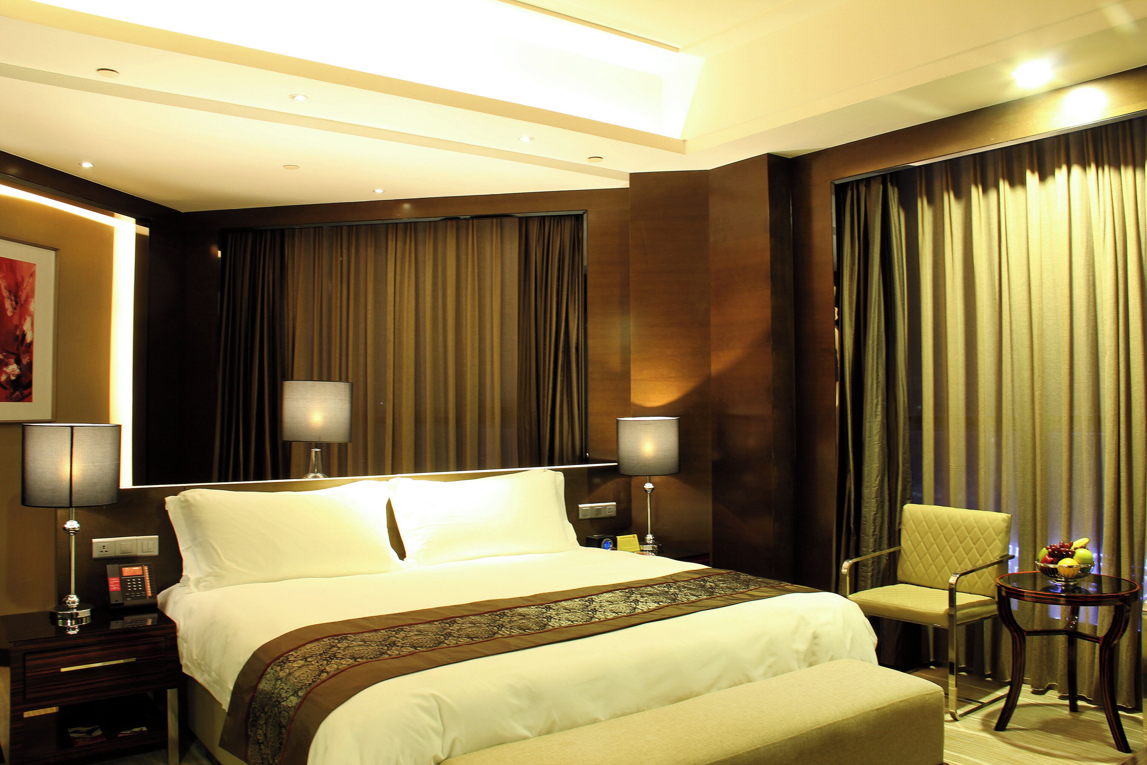 A hotel room with a bed, two bedside lamps, a chair, a small table with fruit, and a painting on the wall.