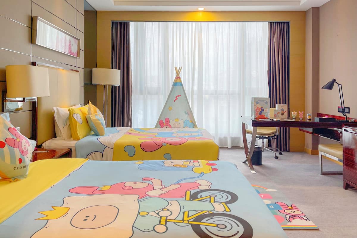 A hotel room with two beds, a desk, a chair, a desk lamp, a window with curtains, and a welcome sign.