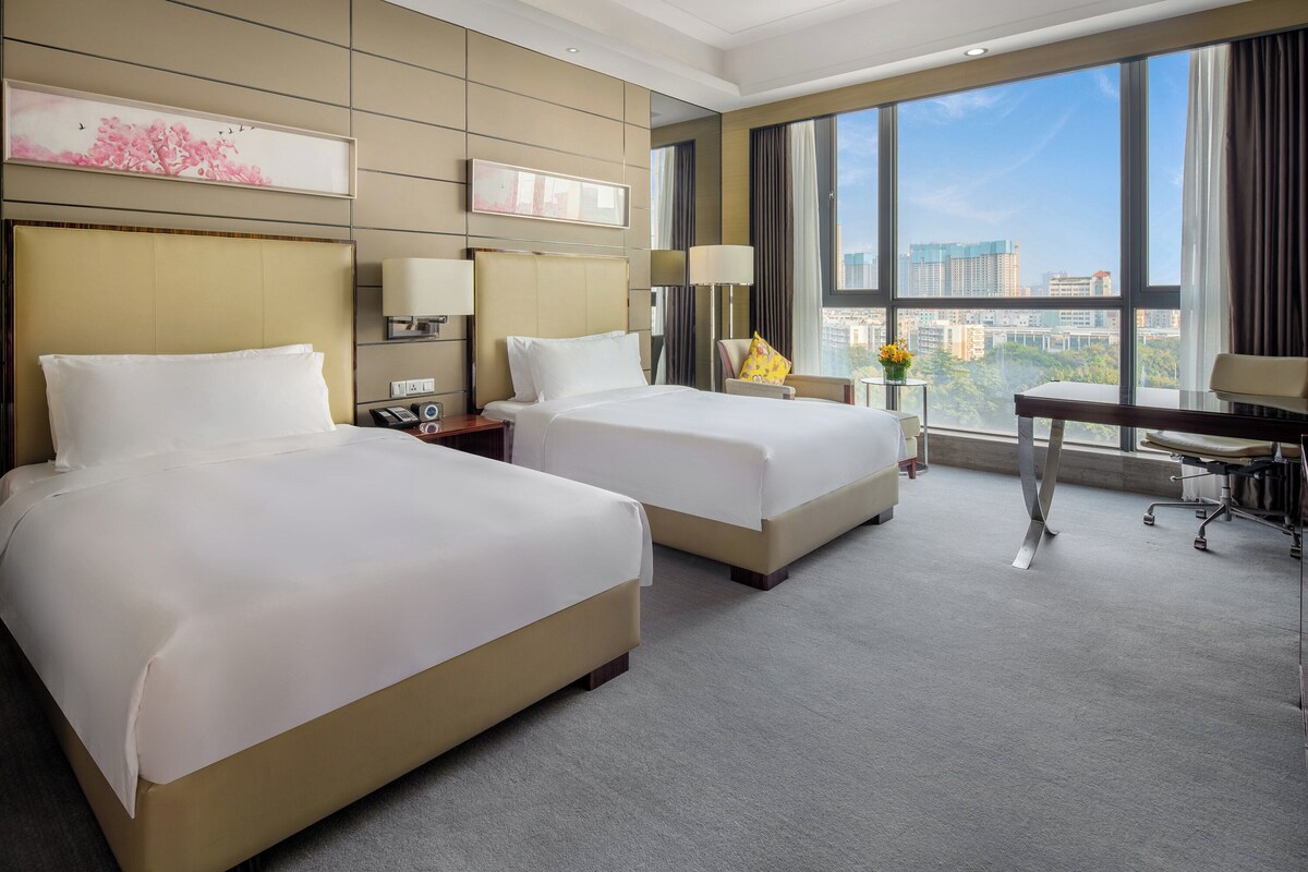 A hotel room with two beds, a desk, a chair, and a large window offering a city view.
