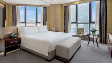 Een moderne hotelkamer met een groot bed, een nachtkastje met een telefoon, een stoel, een klein tafeltje met een vaas, en grote ramen met uitzicht op de stad.