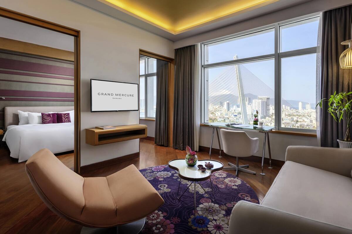 A hotel room with a bed, a desk, a chair, a sofa, a large window with a city view, and a framed sign on the wall.