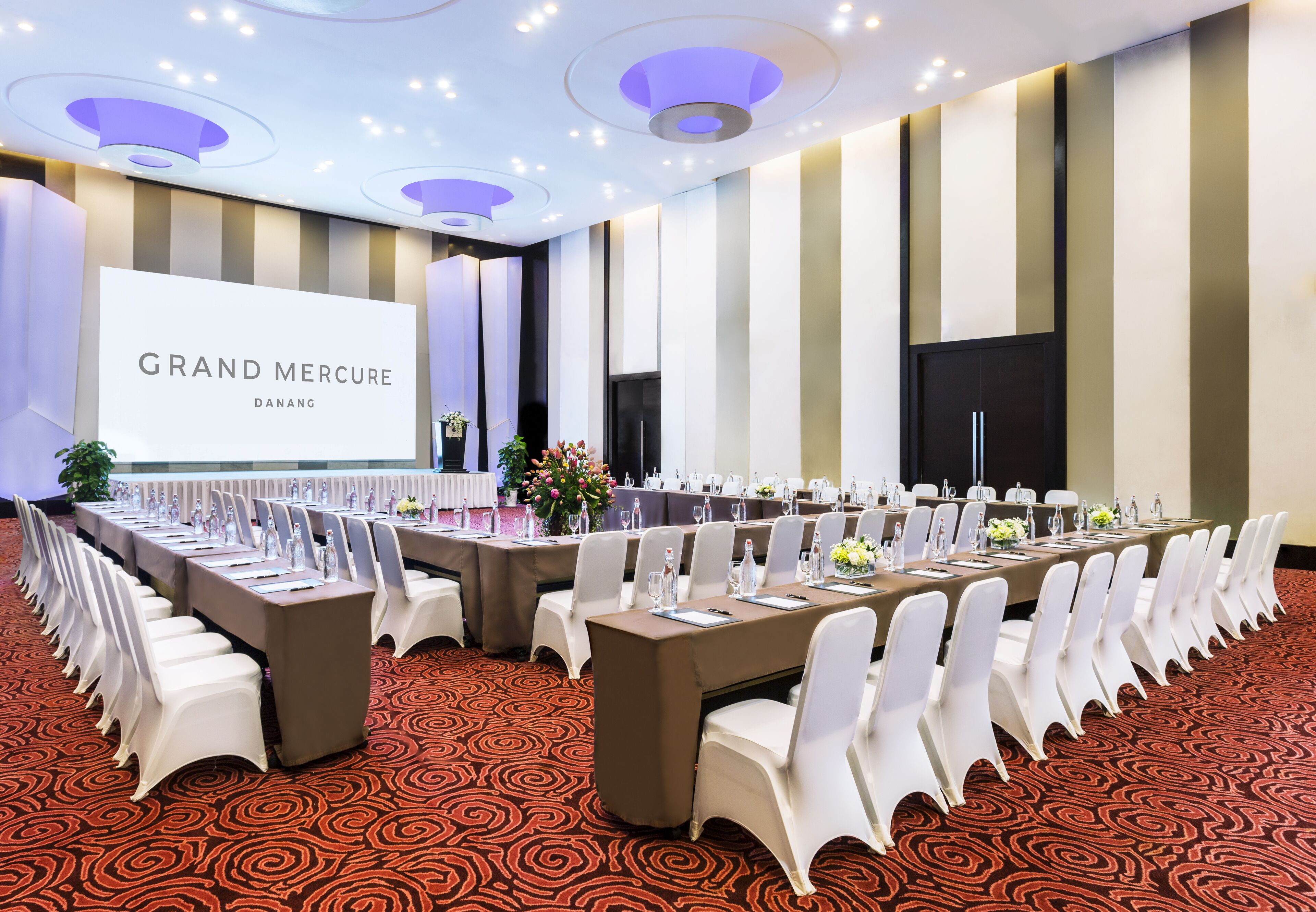 A conference room with a long table, white chairs, and a large screen displaying "GRAND MERCURE DANANG".