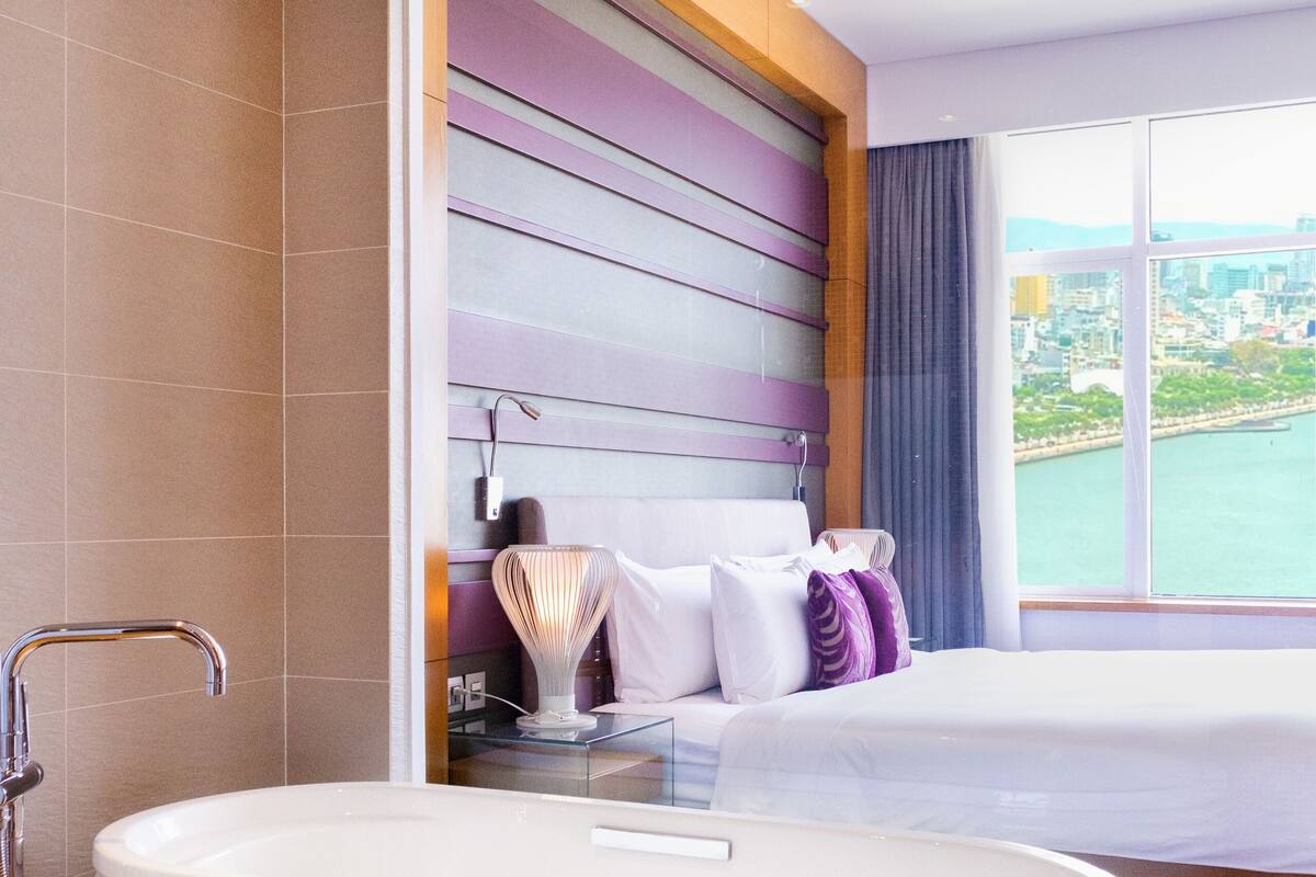 A modern hotel room with a large bathtub, a bed with a view of the city, and a cityscape visible through the window.